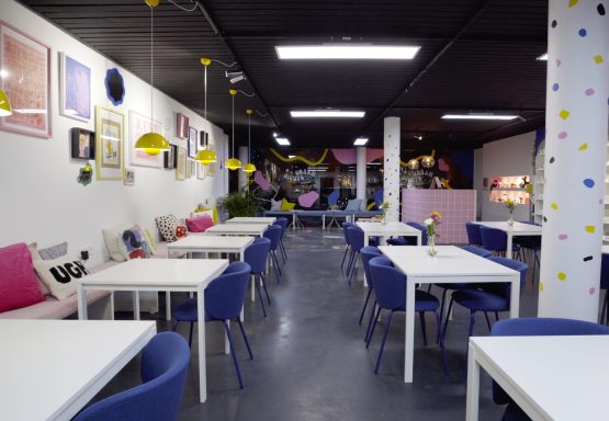 Helle Cafésarena mit Tischen, blauen Stühlen und bunten Dekorationen.