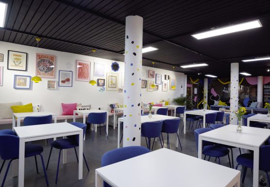 Helle, moderne Café-Atmosphäre mit Tischen, bunten Stühlen und dekorativen Wänden.